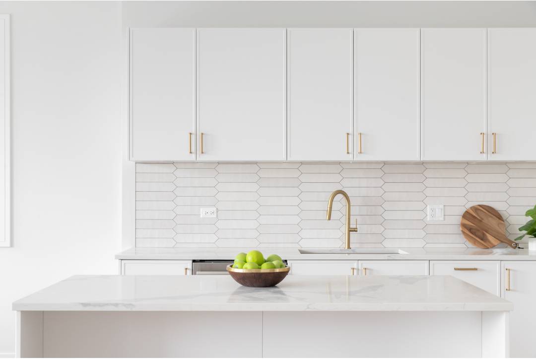An all-white kitchen with white picket subway backsplash tiles and white cabinets with gold accents. The subway backsplash tiles are installed with gray grouting.